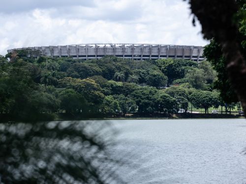  A imagem mostra o estádio Mineirão visto de longe, parcialmente escondido pela vegetação. A parte superior da sua estrutura circular de concreto se destaca acima da densa mata verde. Em primeiro plano, um lago ocupa a metade inferior da foto, refletindo as nuvens do céu. Uma árvore desfocada no lado direito e um arbusto no esquerdo emolduram a cena, sugerindo uma vista do complexo da Pampulha. O céu está nublado, com nuvens pesadas.