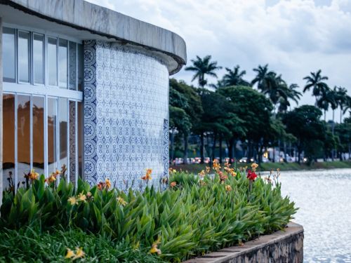 A fachada curva da Casa do Baile de perto, coberta por azulejos brancos e azuis com desenhos florais. Em primeiro plano, há um canteiro com folhas verdes e pequenas flores laranjas e vermelhas. Ao lado, a Lagoa da Pampulha. A outra margem tem uma fileira de palmeiras e carros com faróis acesos.