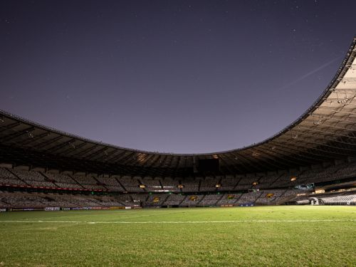  A imagem mostra uma vista noturna do campo do estádio Mineirão. O gramado verde, levemente iluminado, ocupa o primeiro plano. Acima dele, o teto do estádio se curva em arco, cobrindo as arquibancadas com fileiras de cadeiras brancas. No topo, há um vasto céu noturno escuro, pontilhado muitas estrelas. 