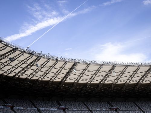  A imagem mostra o teto do estádio Mineirão e parte das arquibancadas sob a luz do dia. A estrutura angular de aço e o material translúcido formam o teto curvo do estádio. As fileiras de cadeiras cinzas e brancas são vistas abaixo, em sombra. O céu, em um tom claro de azul, contrasta com as nuvens finas e brancas, além de uma longa trilha de fumaça, provavelmente de um avião, cruzando a imagem. 