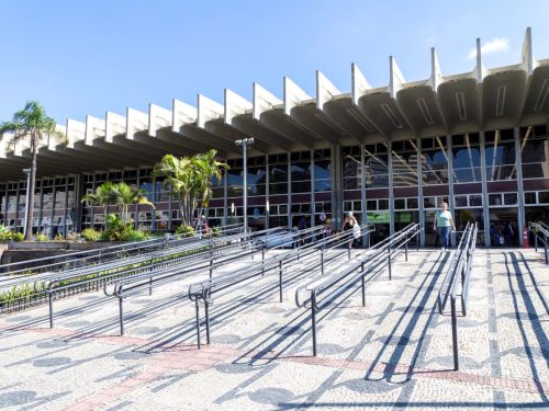 Fachada da rodoviária em um dia de sol. Em primeiro plano, uma calçada com piso de pedras portuguesas e várias rampas de acesso com corrimãos de metal, que projetam longas sombras no chão. Ao fundo, o prédio com sua icônica marquise de concreto com vigas aparentes e uma grande fachada de vidro.