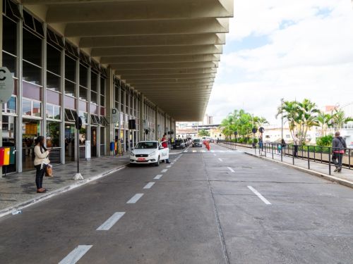 Área externa de embarque e desembarque. À esquerda, a fachada de vidro e os pilares de sustentação do terminal, com placas indicando os portões. A grande marquise de concreto cobre a via. Um carro branco está parado. À direita, uma calçada com grade de proteção e algumas pessoas aguardando.