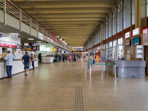 Saguão interno e amplo da rodoviária, com piso bege e teto com longas vigas. À esquerda, guichês de venda de passagens com letreiros luminosos e algumas pessoas na fila. À direita, um balcão de informações de granito e uma parede de vidro que vai do chão ao teto, mostrando a área externa.
