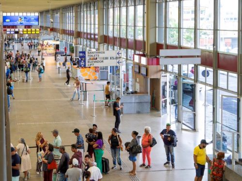 Vista de um mezanino do saguão principal, mostrando o movimento de passageiros. As pessoas formam filas nos guichês, caminham com malas ou aguardam em grupos. À direita, uma parede de vidro do chão ao teto ilumina o ambiente. O piso bege tem faixas de sinalização tátil.