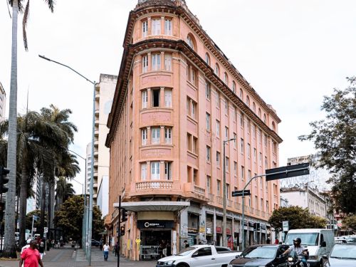 Edifício histórico rosa, de esquina, com quatro andares, janelas retangulares e cúpula arredondada no topo. No térreo há vitrines comerciais. Em frente, faixa de pedestres larga em vermelho e branco, carros parados e postes de sinalização.