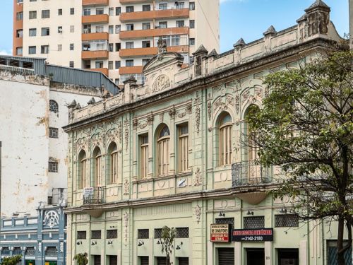 Prédio antigo verde-claro de dois andares, com janelas arqueadas, detalhes em relevo e cornijas brancas. Ao lado, edifícios modernos mais altos, em tons bege e laranja, contrastando com a arquitetura histórica.