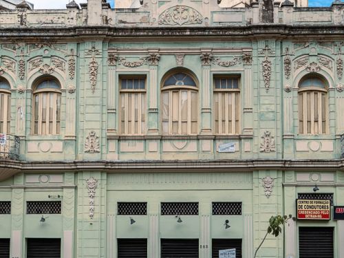 Fachada de prédio verde-claro de dois pavimentos com portas metálicas fechadas no térreo e janelas altas em arco no andar superior. Um homem de mochila caminha na calçada, e a rua aparece parcialmente.