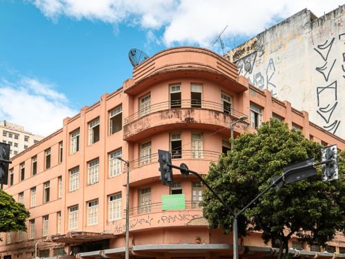 Prédio rosa de esquina com três andares, sacada curva com grades no segundo piso e fachada ornamentada. No térreo, lojas comerciais abertas e letreiros. Pedestres atravessam a faixa de pedestres em frente.
