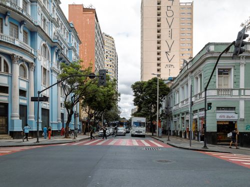 Cruzamento com faixa larga em vermelho e branco. À esquerda, prédio azul de fachada clássica com colunas e sacadas; à direita, prédio verde-claro baixo com portas metálicas. Ao fundo, prédios modernos mais altos.