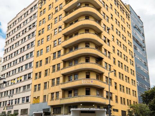 Edifício amarelo de esquina, com vários andares, sacadas curvas e janelas retangulares alinhadas. No térreo funcionam lojas com letreiros visíveis. Há pedestres caminhando e postes de sinalização próximos.