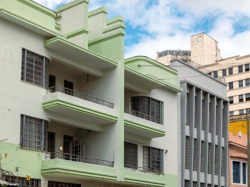 Prédio verde-claro de quatro pavimentos, com sacadas de vidro arredondadas e fachada simples. Em volta, calçadas largas com árvores. Ao fundo, torres residenciais altas em tons de bege e amarelo.