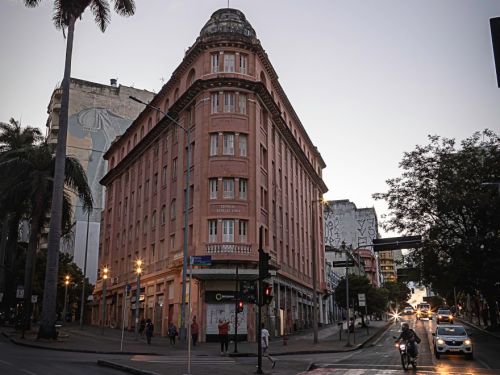 Edifício rosa de esquina, mesmo da primeira imagem, visto ao entardecer. A cúpula e sacadas estão iluminadas por postes de luz e faróis de carros. Pedestres atravessam a rua e há movimento intenso no cruzamento.