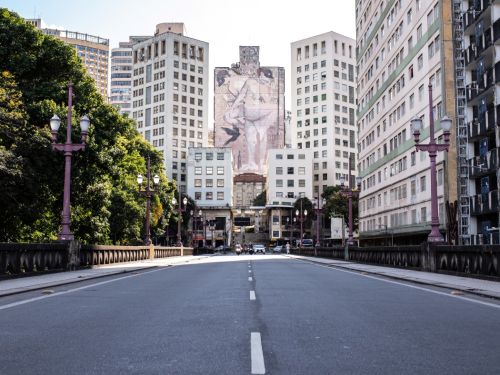 A pista de asfalto do viaduto é vista de frente, em perspectiva, conduzindo o olhar para o centro da cidade. Nas laterais, há postes de iluminação de cor vinho. Ao fundo, um grande mural de arte urbana com figuras humanas se destaca na fachada de um dos altos edifícios que formam um corredor.