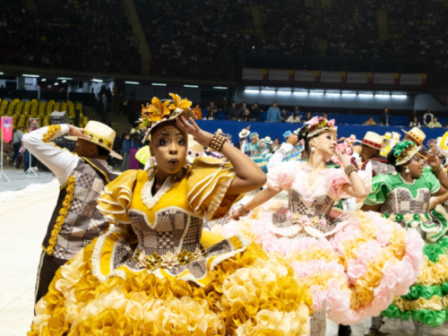 Um grupo de quadrilha junina se apresentando em um tablado, com destaque para uma mulher de vestido amarelo a frente da foto.