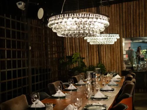Foto da ambientação interna do restaurante Spicy, mostrando uma grande mesa de madeira posta com taças e pratos e dois grandes lustres de teto centralizados a mesa 