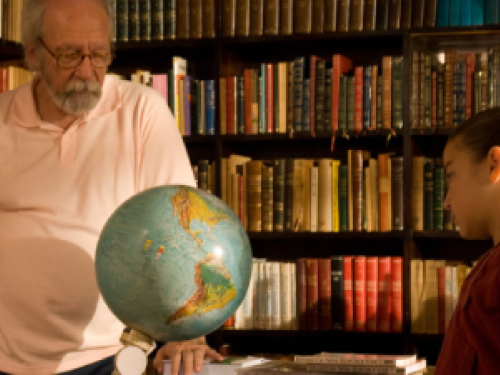 Foto no cenário de uma biblioteca, com duas pessoas, um senhor e uma menina olhando para o globo