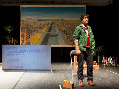 Foto de ator com feição de riste com um com um mapa de uma cidade, com um córrego central, estirada em uma bandeira atrás dele. 