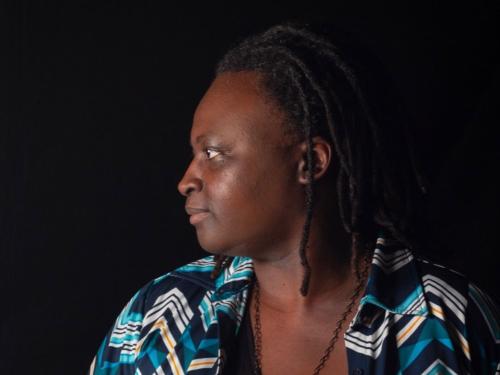 Mulher negra com cabelos soltos. Ela está de perfil, olhando para o lado esquerdo. Usa uma camisa preta e um casaco colorido em tons de azul e verde