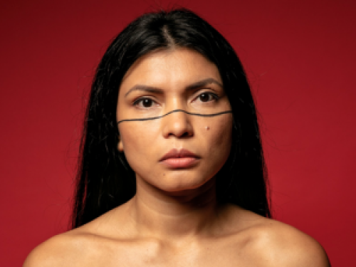 na foto, uma mulher indígena parda de cabelos lisos e pretos. Seu rosto tem uma pintura com uma linha preta marcando pela metade o rosto, abaixo dos olhos em todo o rosto.