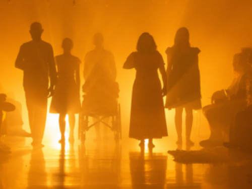 Foto com uma luz no fundo amarelada mostra os corpos dos atores em silhuetas