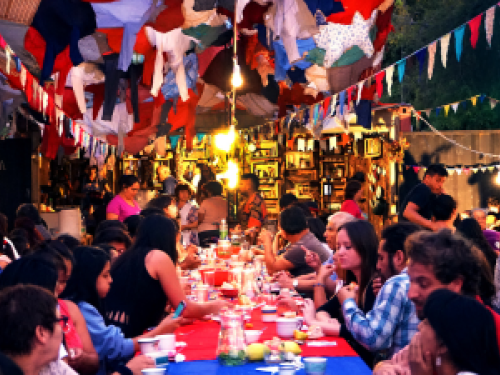 Na foto, pessoas compartilham comidas em uma mesa comunitária