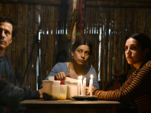 Uma Família chilena sentada à mesa, na luz de velas, se entreolham de forma tensa. Estão sentados à mesa, duas mulheres e um homem.