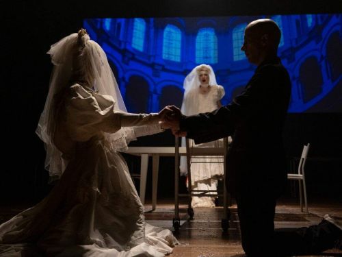Na foto, um casal ajoelhado segura as mãos, a noiva com véu, enquanto ao fundo uma outra noiva rege o casamento.