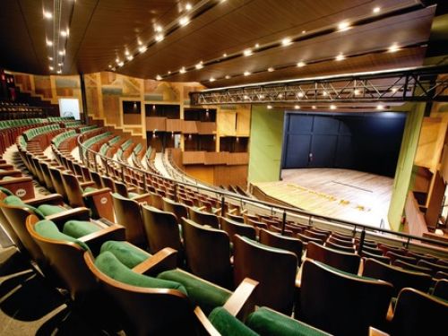 Área interna do teatro, vista do ponto superior para o primeiro andar e palco, ambiente bem iluminado e com bancos verdes e assentos almofadados 