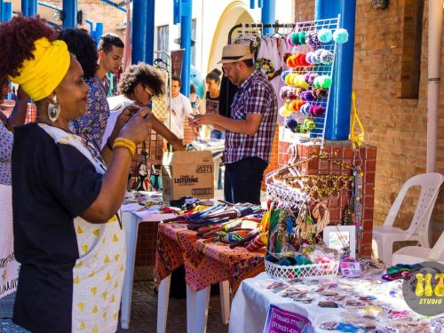 Foto de espaço com pessoas ao fundo e mesas de exposição de acessórios e adereços afro. 