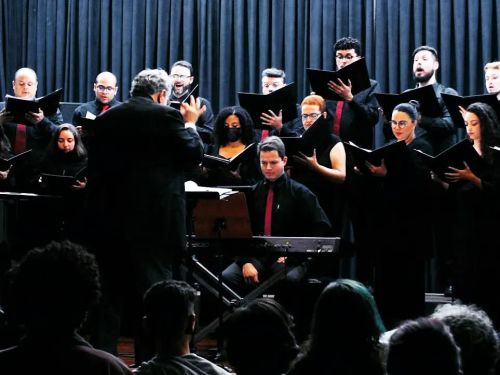A imagem mostra um coral com várias pessoas em pé, todas vestindo preto, em um palco. 