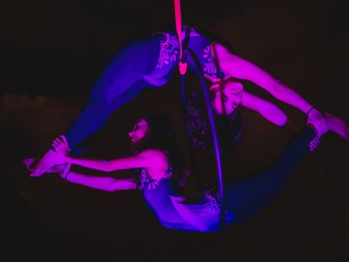 Foto de duas pessoas suspendidas por um bambole, com luzes roxas e fundo preto