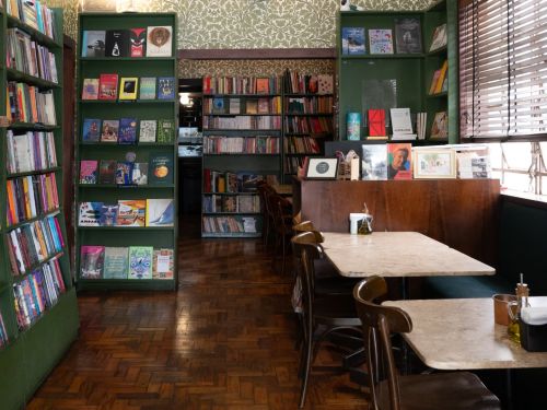 Foto do interior do estabelecimento, com estantes de livros, piso de taco e mesas.