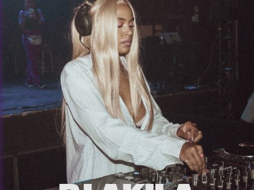 DJ Akila com fones grandes e cabelo longo e claro está tocando em uma mesa de som, em um ambiente escuro de show. O texto anuncia “DJ Akila” e o show “Budah”.