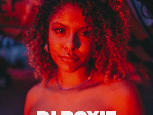 Uma DJ de cabelo cacheado e iluminado em tons vermelhos olha para a câmera em um ambiente noturno. O texto destaca “DJ Roxie” e o show “Budah”.