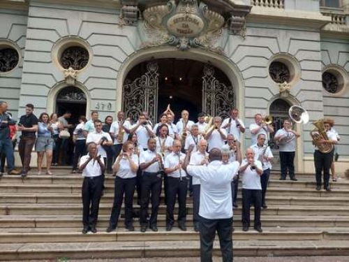 A foto mostra uma banda músicos e instrumentistas vestidos de branco nas escadas da secretaria da aviação.