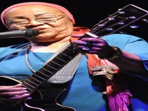 Imagem real de dona Jandira segurando um violão e cantando 