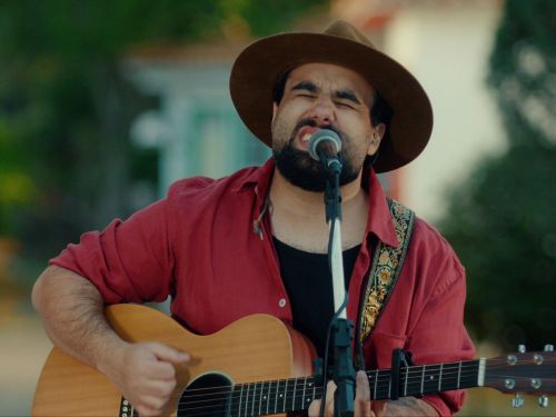 Um homem toca violão e canta ao ar livre. Ele usa chapéu marrom, camisa vermelha e toca diante de um microfone. Atrás dele há casas antigas coloridas e árvores.