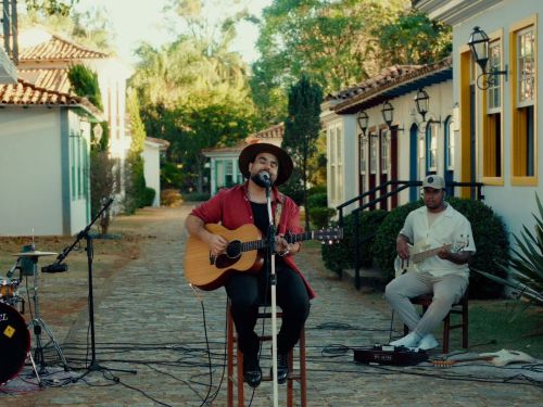 Um homem toca violão e canta ao ar livre. Ele usa chapéu marrom, camisa vermelha e toca diante de um microfone. Atrás dele há casas antigas coloridas e árvores.