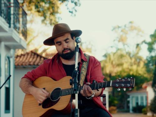 Um homem toca violão e canta ao ar livre. Ele usa chapéu marrom, camisa vermelha e toca diante de um microfone. Atrás dele há casas antigas coloridas e árvores.