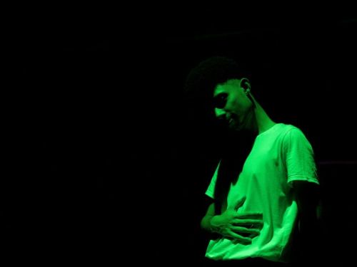 A foto mostra uma pessoa em um ambiente escuro, iluminada por uma luz verde. Ela está usando uma camiseta clara e aparece com uma das mãos sobre o abdômen. O fundo é totalmente preto, destacando apenas o corpo e o rosto sob a iluminação verde.