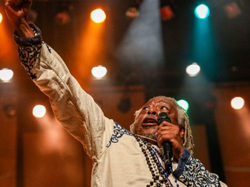 Reprodução sympla Fotografia do artista Mauricio Tizumba no placo com microfone na mão e holofotes ao fundo