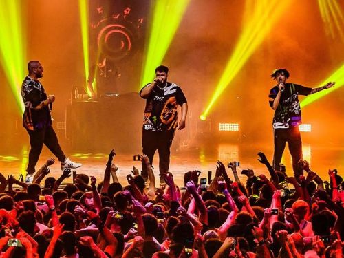 Imagem de um show, com três pessoas cantando.