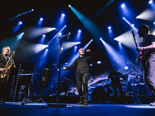 Imagem de um show, com uma banda tocando e um homem em destaque com um microfone na sua frente.