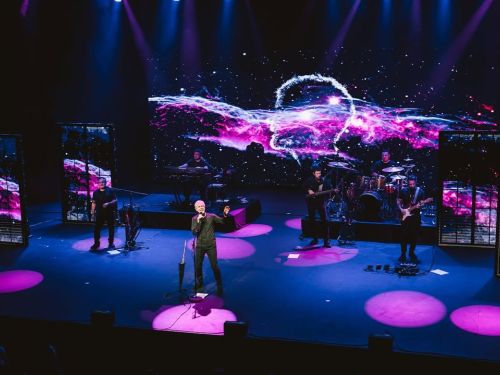 Imagem de um show, com uma banda tocando e um homem em destaque com um microfone na sua frente.