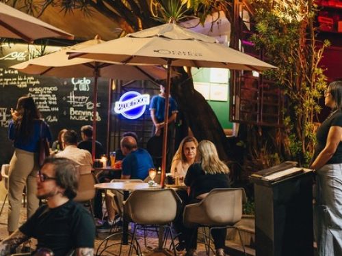 Foto do espaço externo, com mesas, cadeiras, guarda sol, e pessoas ocupando o espaço.