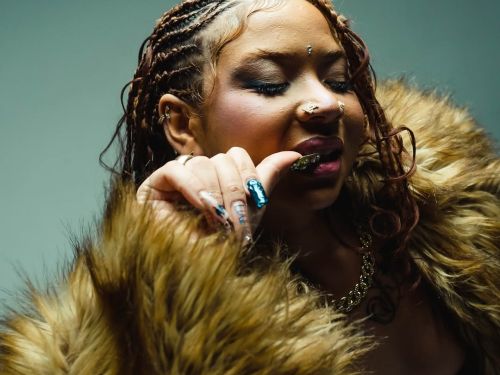 Imagem com uma mulher com a mão na boca, esta usando cum casaco de pelos e os cabelos trançados 