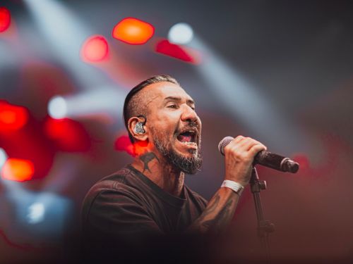 Imagem do cantor Rodolfo Abrantes no palco com microfone, fundo de luzes e holofotes vermelho