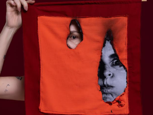 Imagem de uma pessoa segurando em lenço de cor laranja e vermelho, com um buraco no meio e um olho no meio