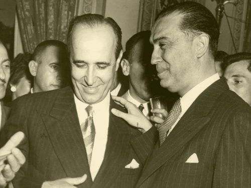 Imagem em preto e branco de dois homens em destaque, um com a cabeça baixa e sorrindo e o outro com as mãos no ombro, sorrindo também