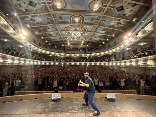Imagem de um teatro, com um homem no palco e uma plateia lotada.